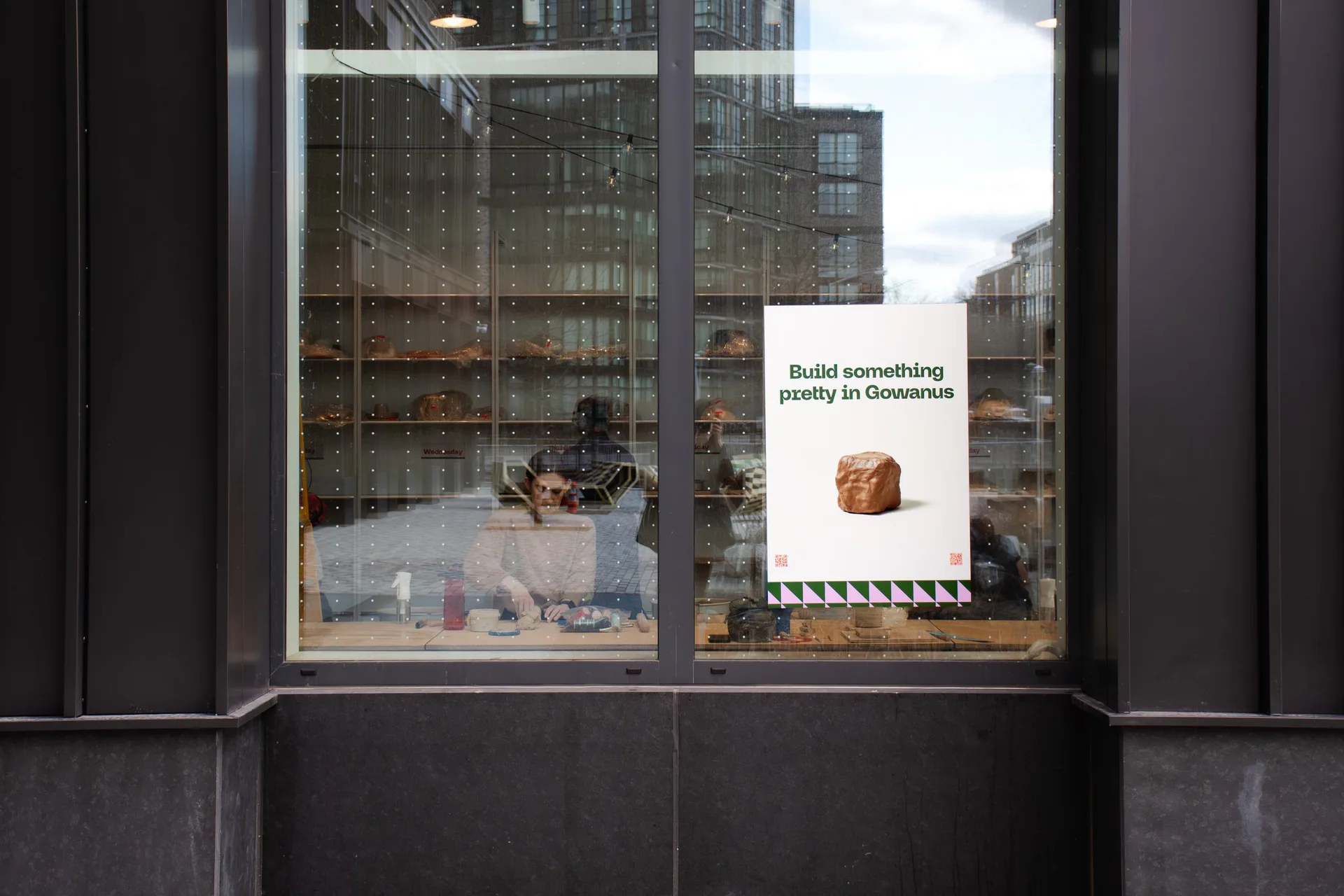 Street-facing storefront windows with city buildings reflected, featuring a white envelope-style sign reading 'Build something costly in Gowanus' with a terracotta vessel image.