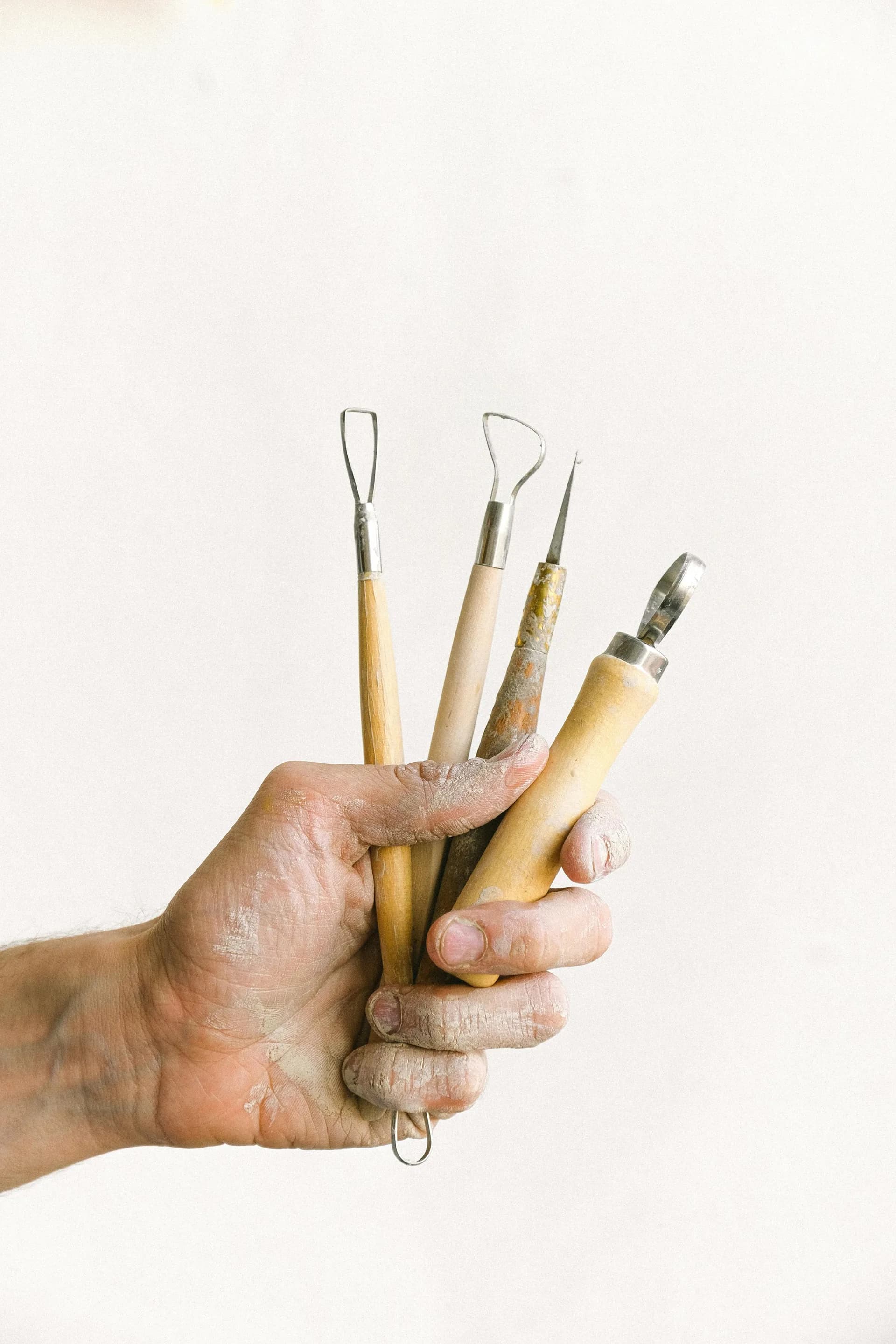 Hand holding five ceramic sculpting tools with wooden and bone handles against a cream background.