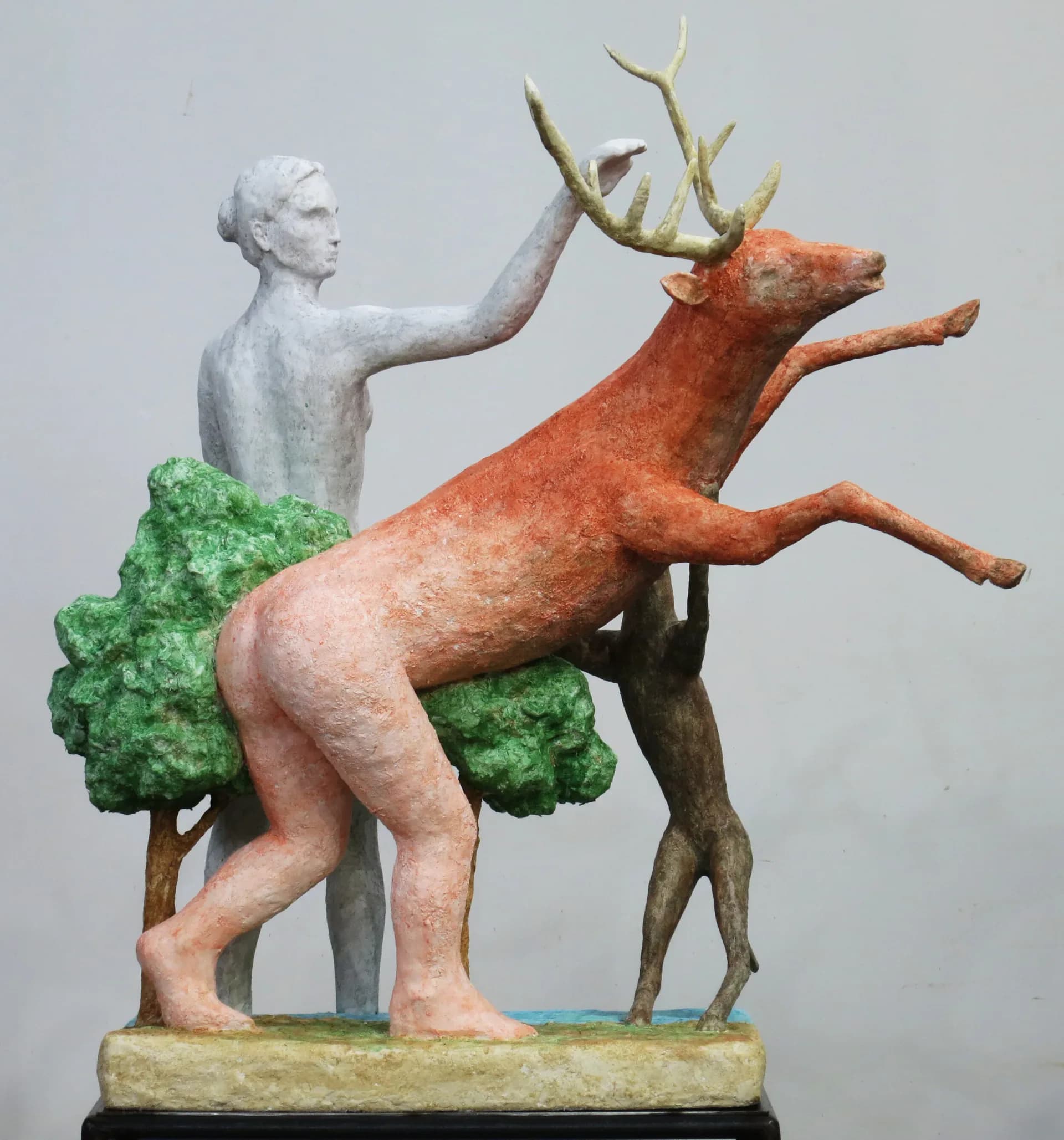 Painted sculpture of a white female figure restraining a rearing red stag with green trees