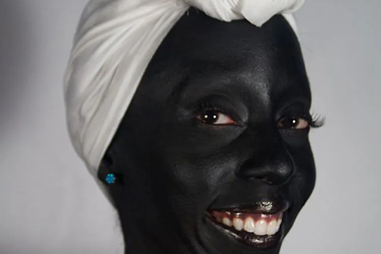 Close-up portrait photograph of a person with darkened face paint, white head wrap, bright pink lipstick, and blue earring, smiling directly at camera against gray background.