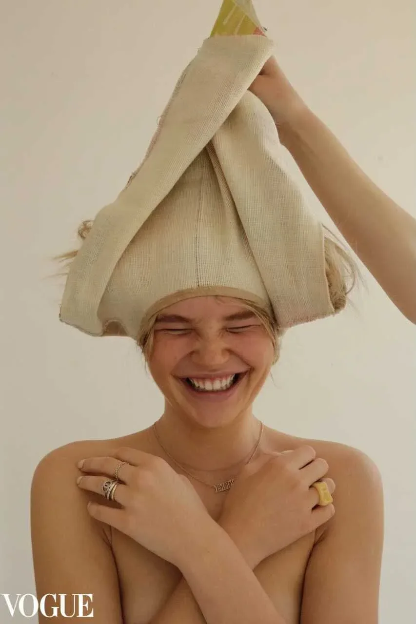 Person smiling with eyes closed, wrapped in cream-colored fabric forming a head covering, wearing rings and a necklace, shot from shoulders up.
