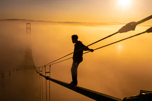 Silhouette of a figure standing on a bridge cable at golden sunrise with fog over water