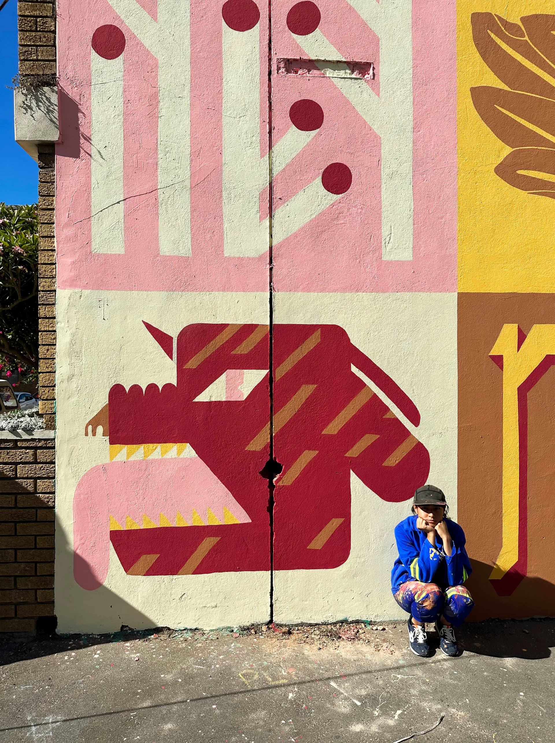 Outdoor mural featuring bold geometric and animal motifs in pink, maroon, and gold on building wall