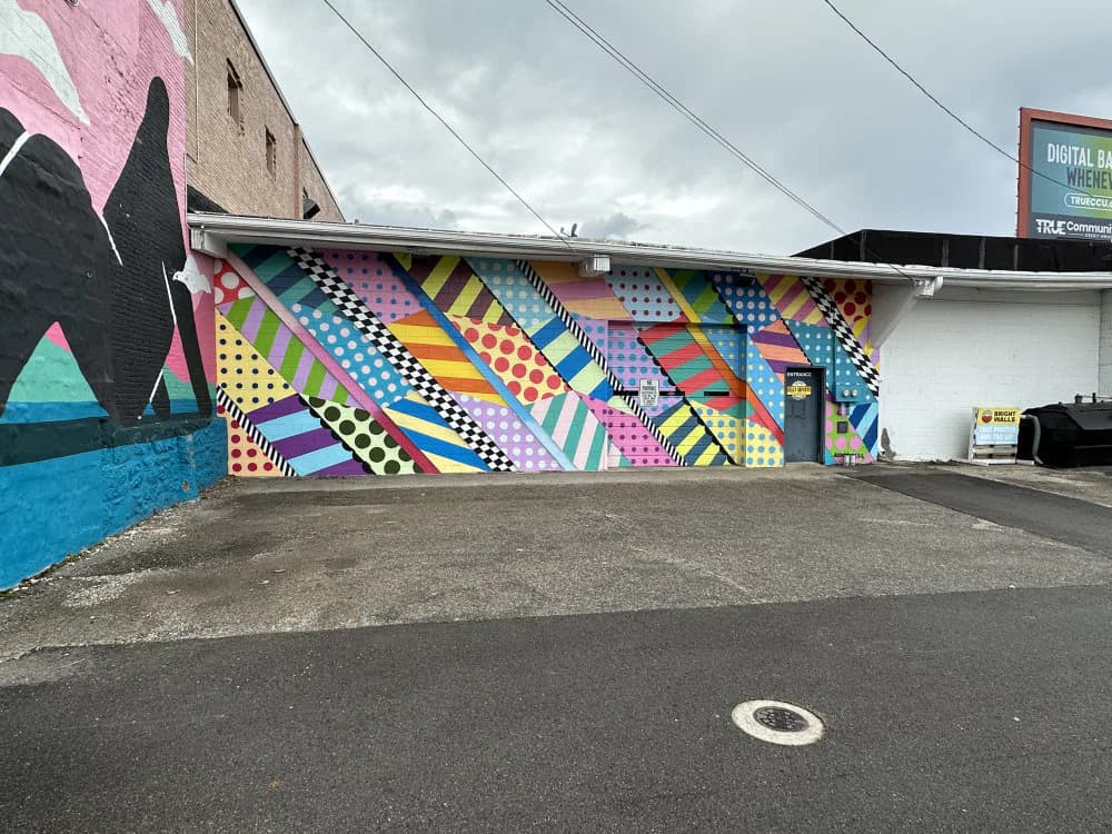 Colorful mural on building wall featuring geometric patterns, stripes, and checkerboard designs in bright blues, yellows, pinks, and purples.