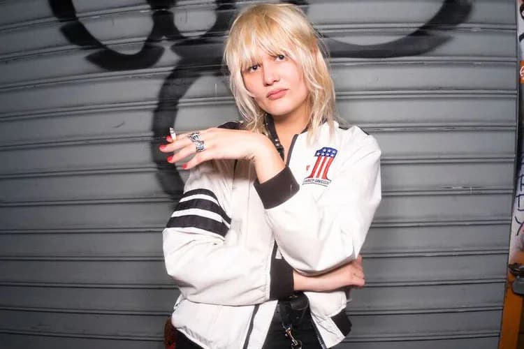 Person with platinum blonde bangs wearing a white bomber jacket with black stripes against a gray metal wall.