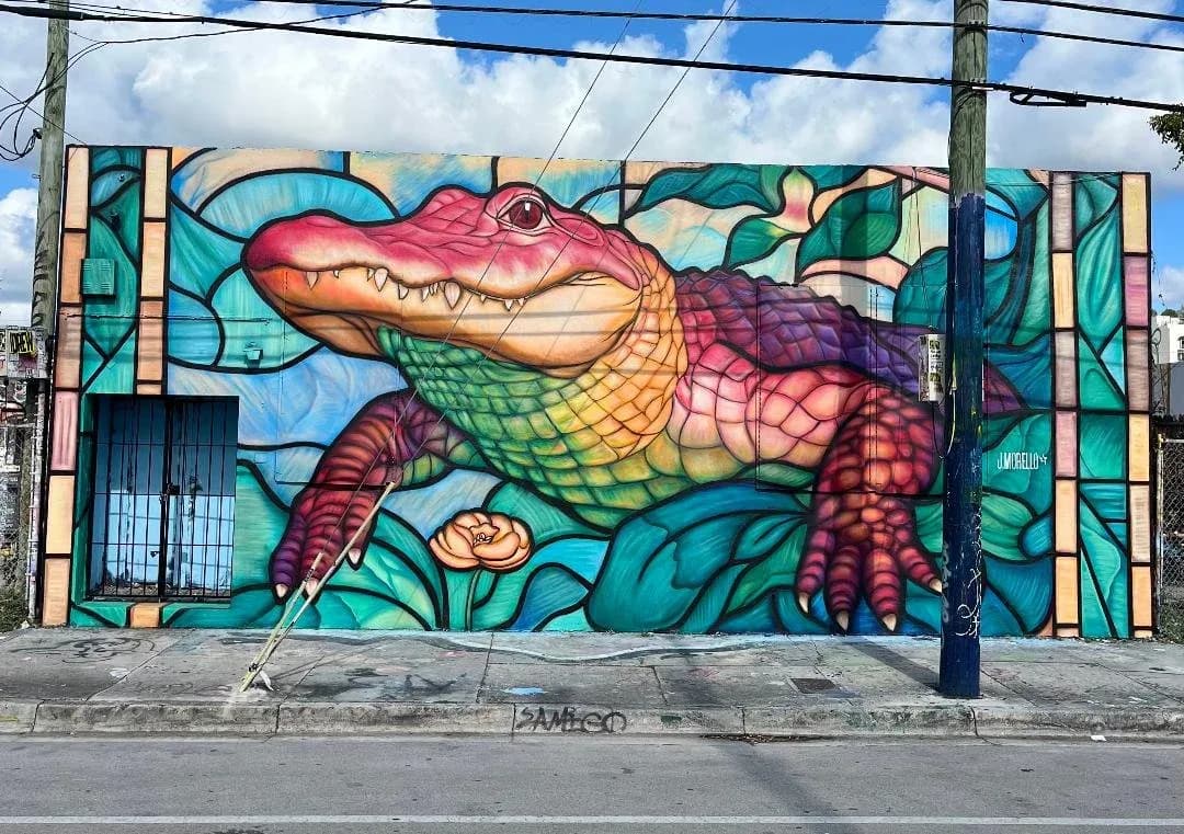 Outdoor mural of rainbow-colored alligator in stained-glass style surrounded by teal foliage and a flower