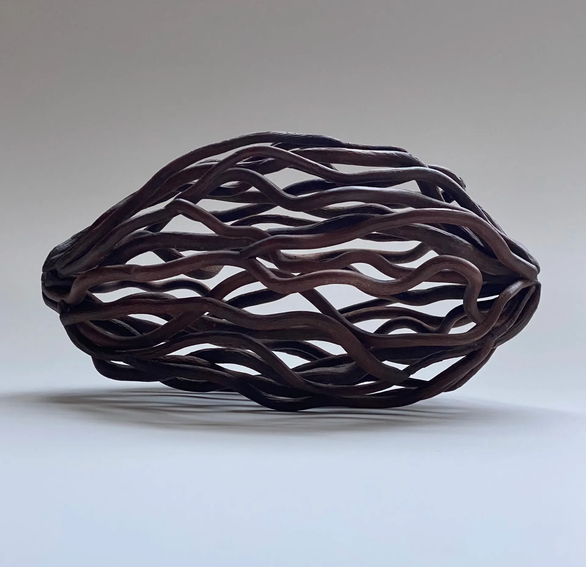 Dark brown woven branch pod sculpture with flowing horizontal wave pattern on neutral background