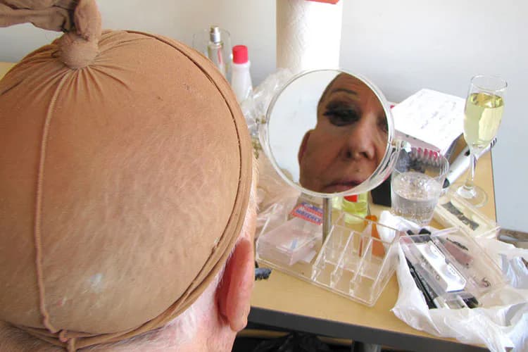 Conceptual self-portrait: person holding circular mirror reflecting their face, surrounded by cosmetic products and bottles on a beige surface.