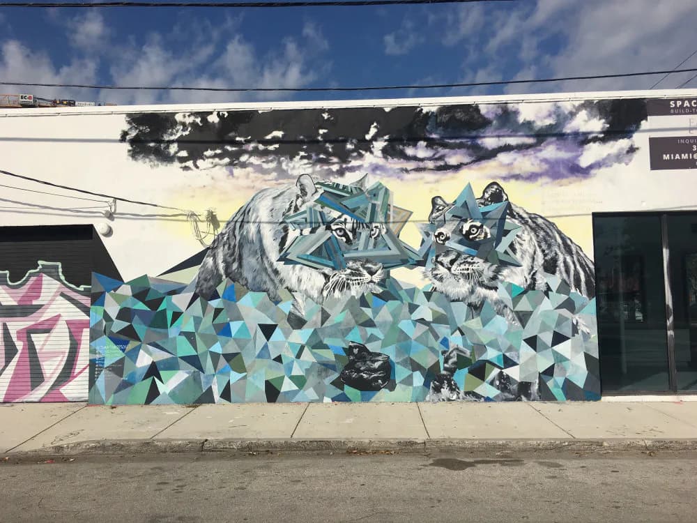 Mural featuring stylized wolf heads in teal and blue tones above geometric triangular patterns, painted on an urban building facade.