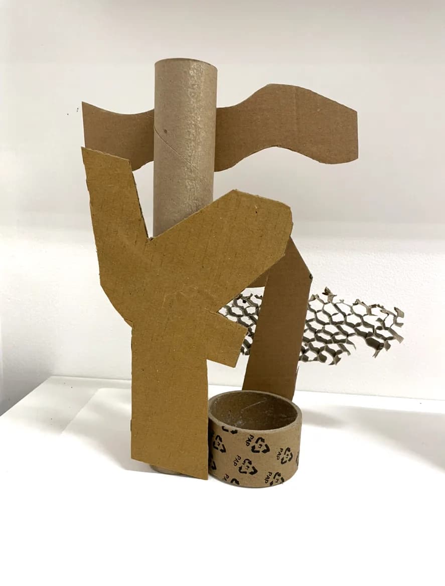 Three-dimensional sculptural assemblage made from cardboard and kraft paper, featuring angular forms, a cylinder, ribbon-like elements, and scattered paper pieces in tan and brown tones.