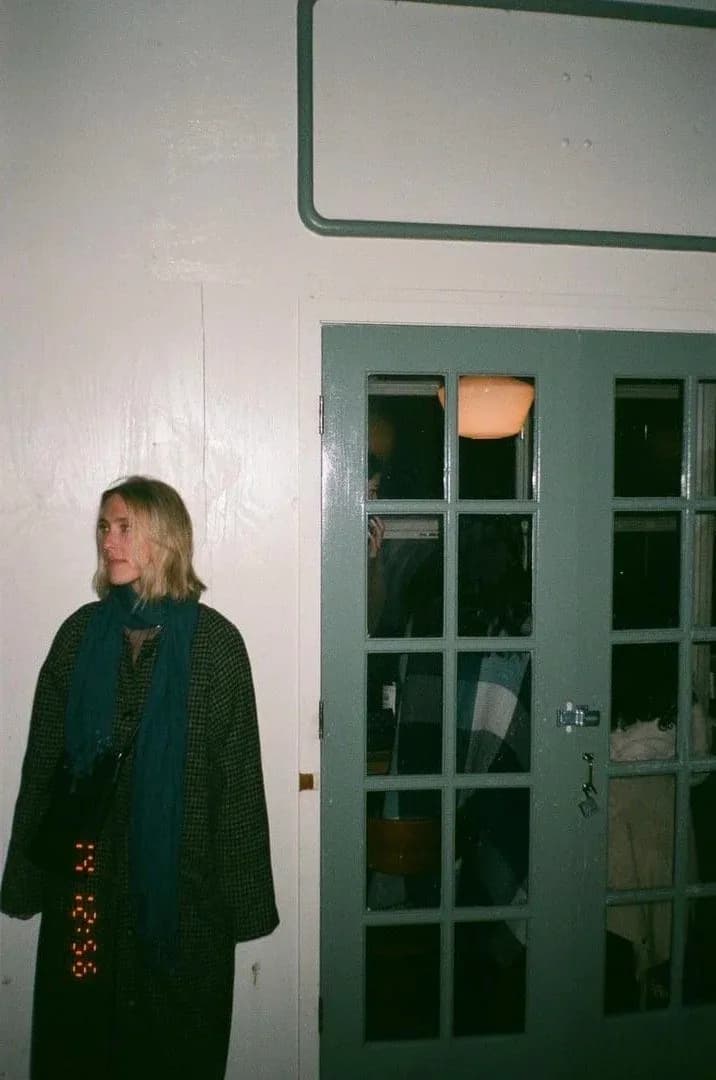Woman with blonde hair in dark clothing standing in an interior gallery space with white walls and a green-framed glass cabinet door.