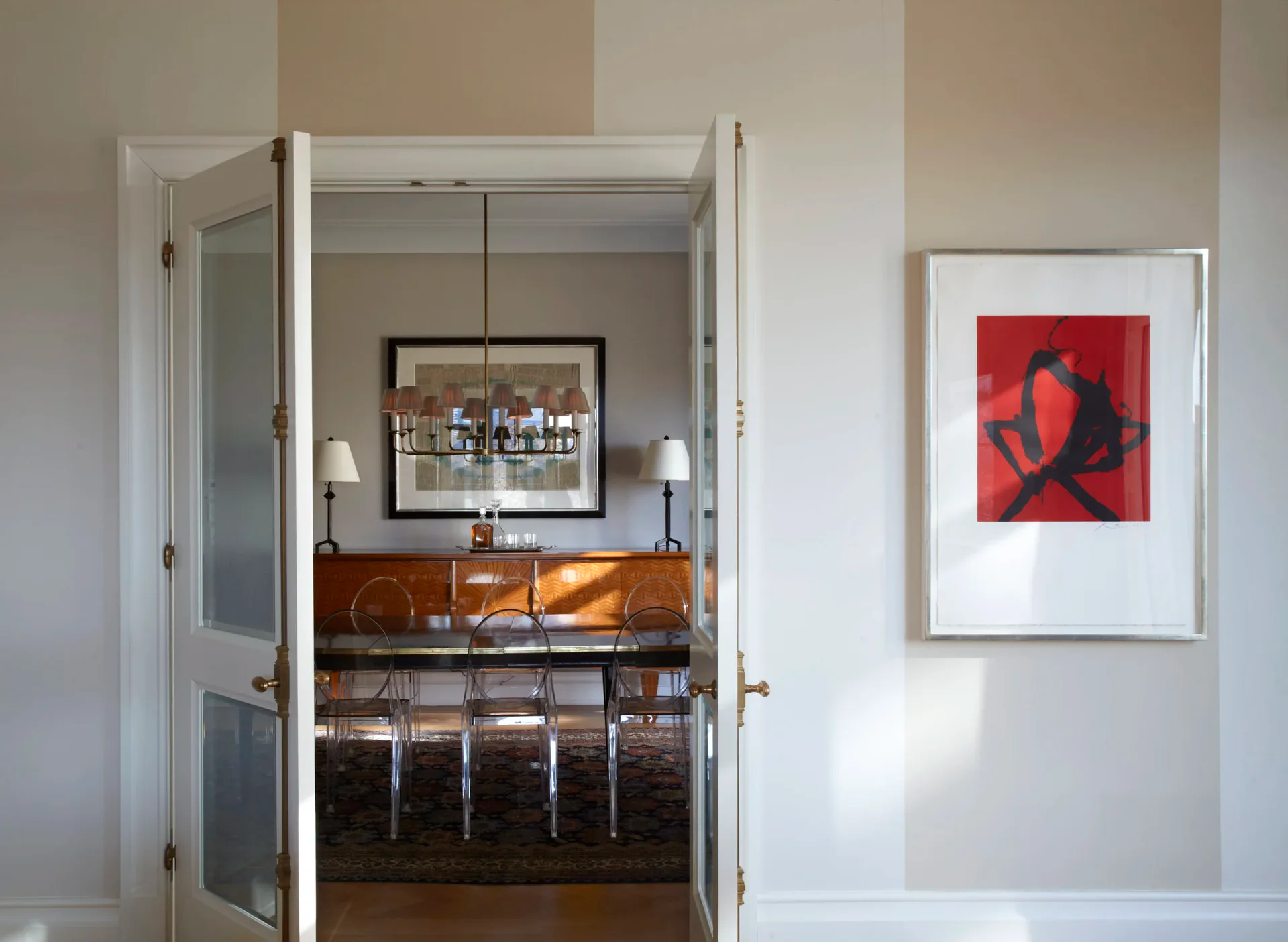 Photograph of a bright gallery interior with white walls, open doorway revealing wooden furniture and bar seating, and a red abstract artwork on the wall.
