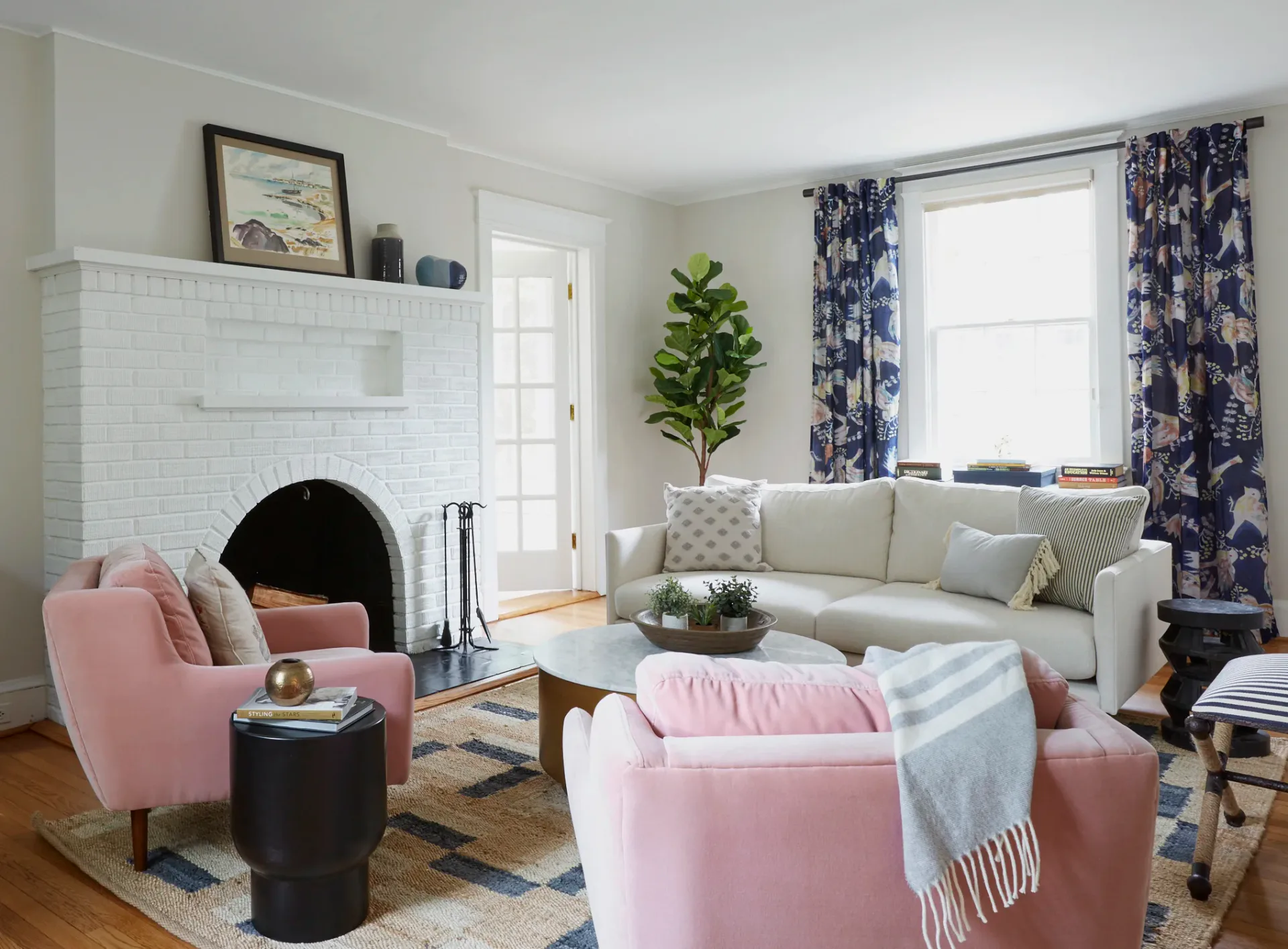 Photograph of a bright living room interior with white fireplace, pink armchairs, patterned curtains, sofa, and wood flooring.