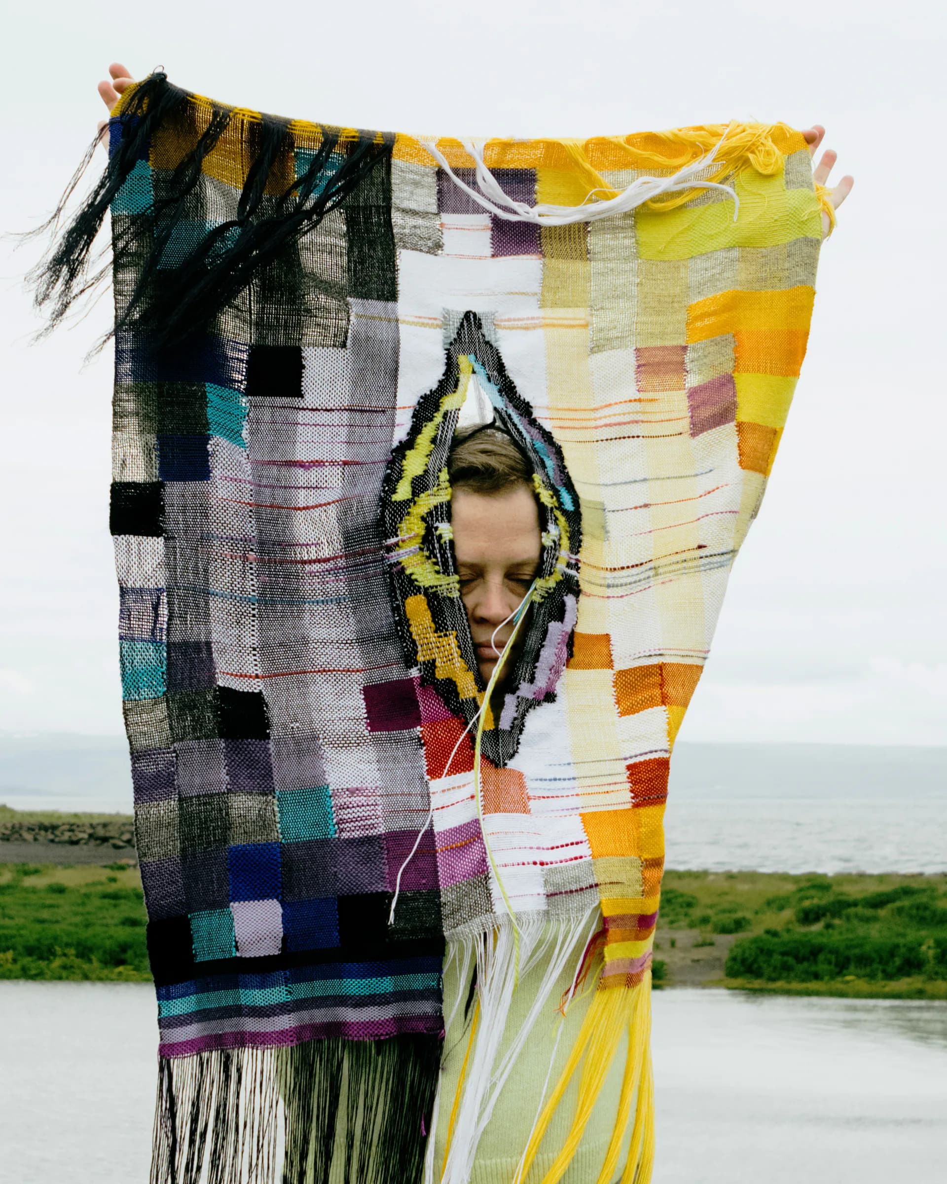 Person holding a large woven textile in multicolored patchwork pattern with yellow, black, and colorful squares, outdoors with water and landscape behind.