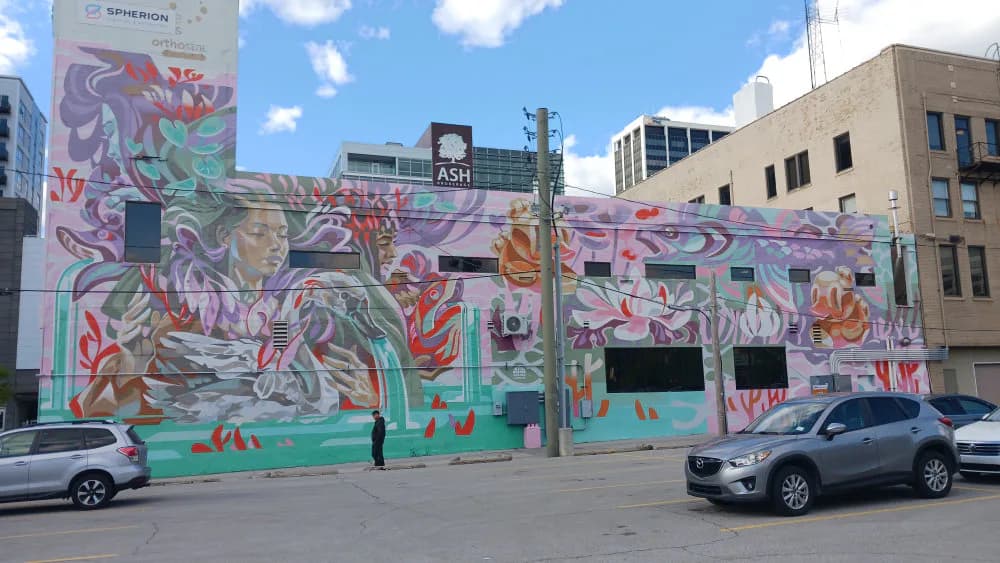 Large-scale mural with vibrant abstract botanical and figurative forms in pink, purple, teal, and orange on a building facade in an urban setting.
