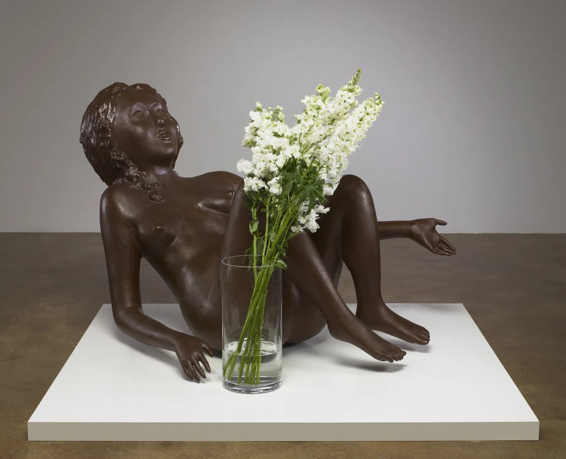 Brown bronze sculpture of a reclining female figure with white flowers in a glass vase placed between her legs on white platform.