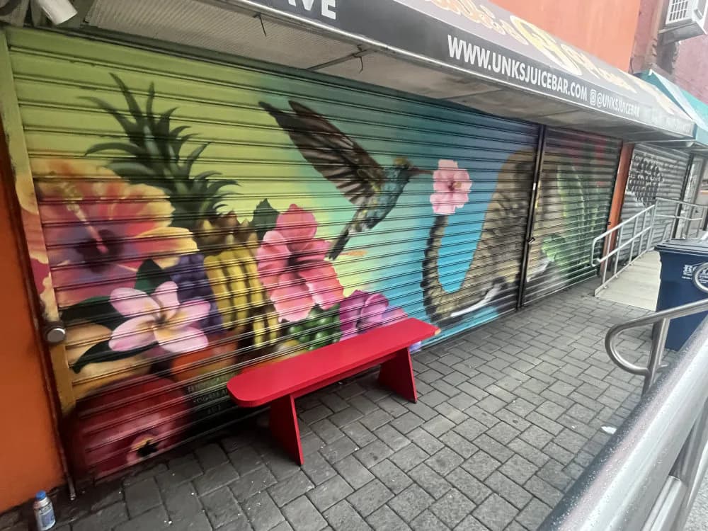 Colorful mural on a storefront shutter depicting a bee pollinating pink flowers with green leaves and blue sky background.