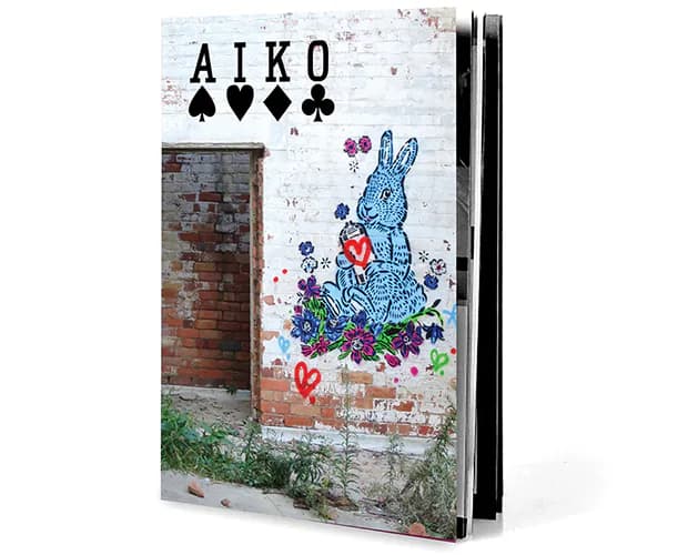 Mixed-media piece on weathered brick wall featuring 'AIKO' logo and blue stenciled rabbit surrounded by colorful floral elements.