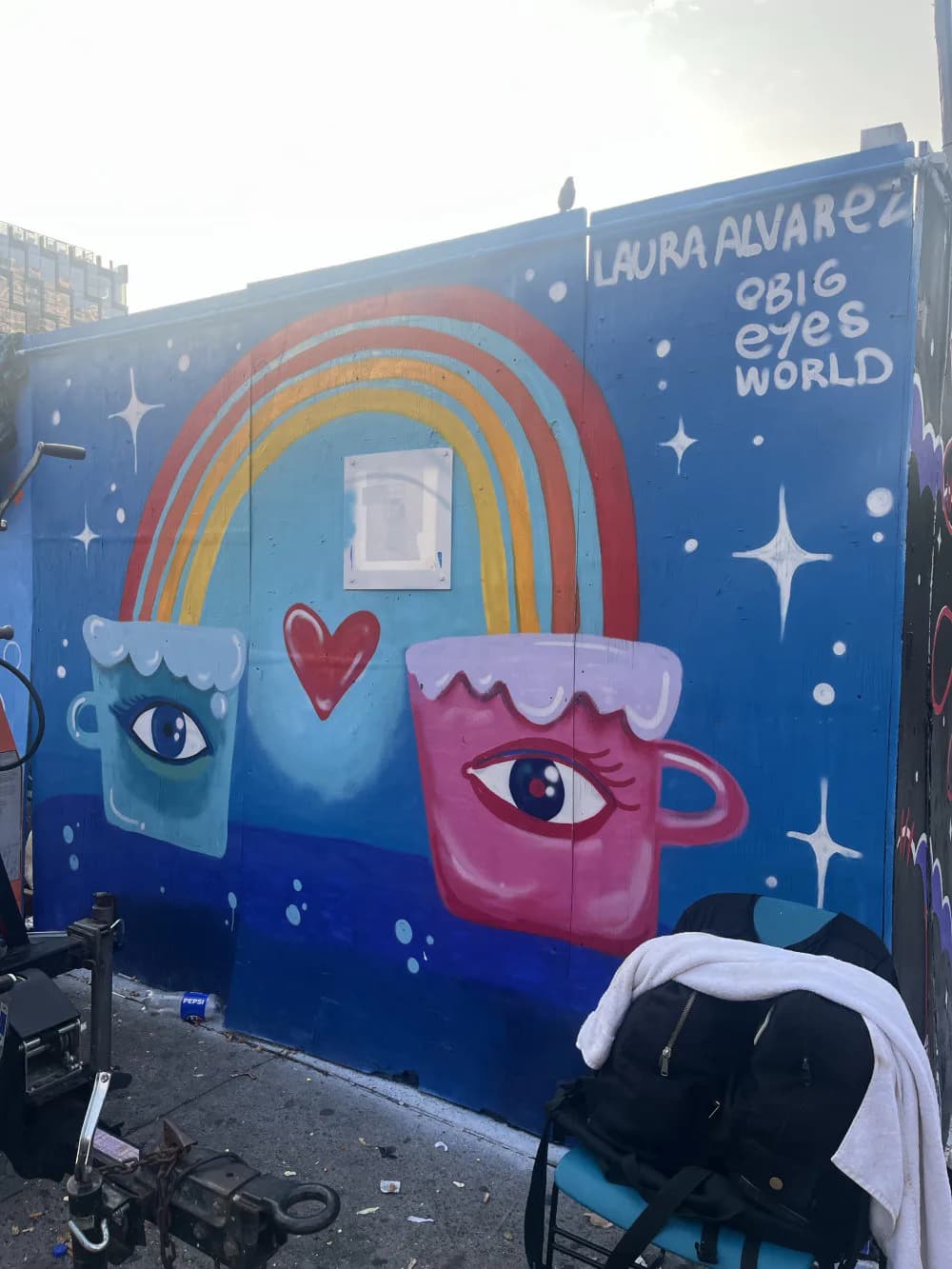 Mural by Laura Alvarez featuring two stylized mugs with eyes and a heart between them, connected by a rainbow on a deep blue starry background.
