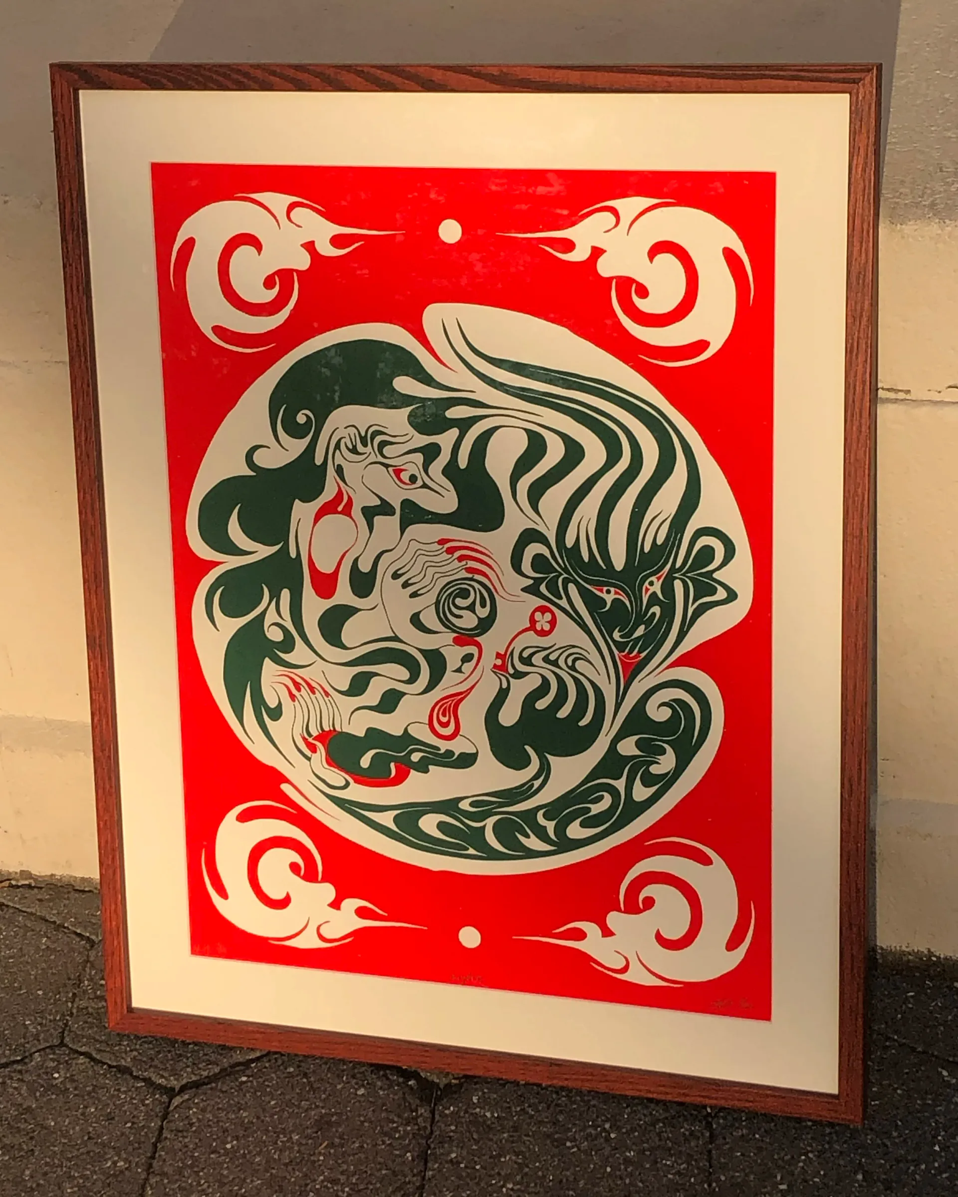 Traditional red and cream silkscreen-style print featuring a central circular creature face with green and black linework, surrounded by ornamental corner spirals in a framed presentation.