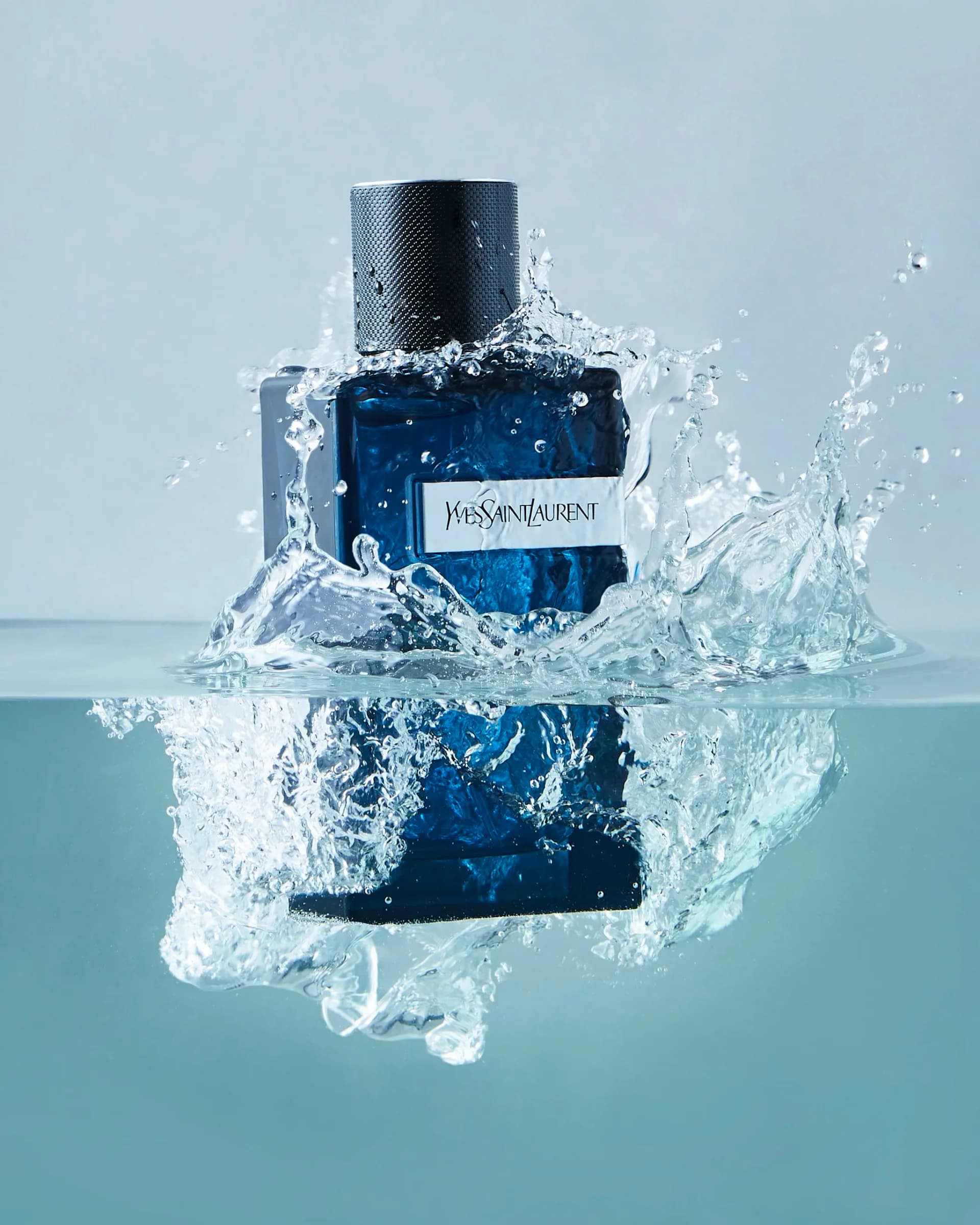 Product photography of a deep blue Yves Saint Laurent fragrance bottle splashing in water against light blue background.
