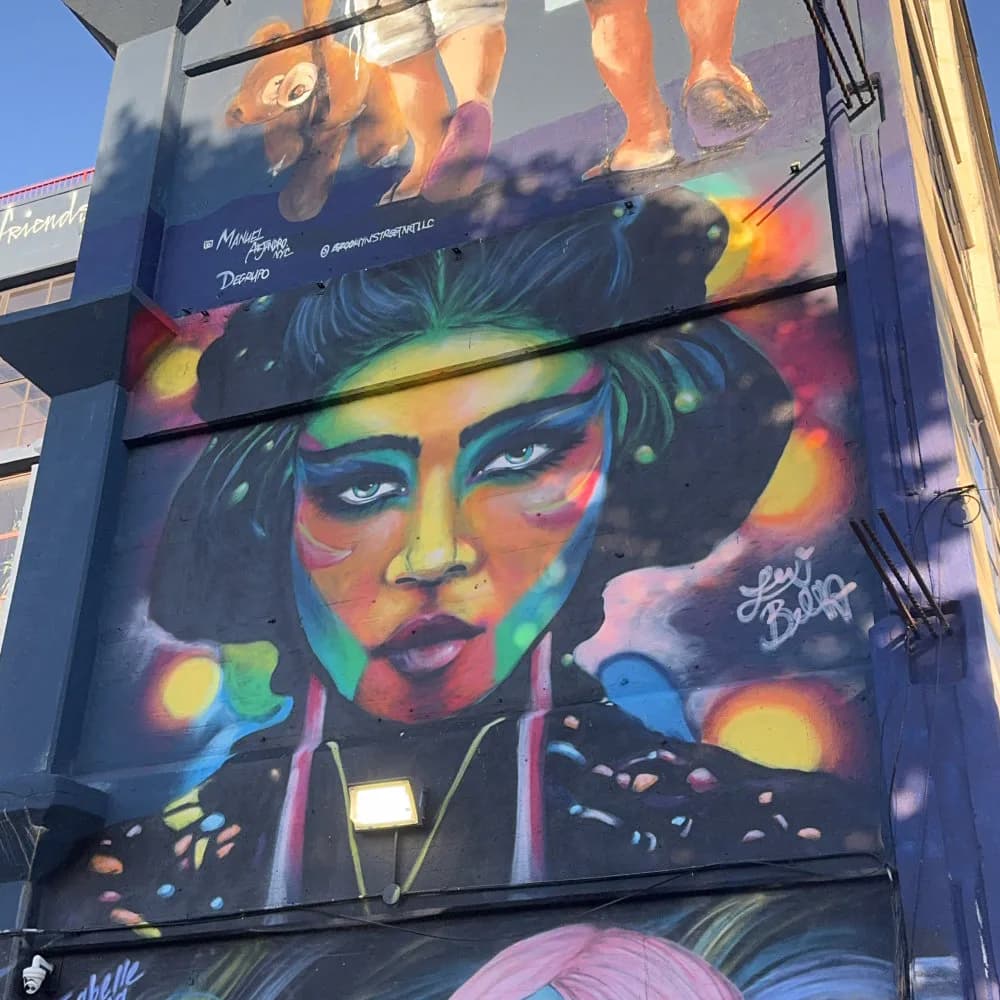 Vibrant street mural depicting a person's face with rainbow-colored geometric patterns, neon accents, and expressive eyes against a dark background.