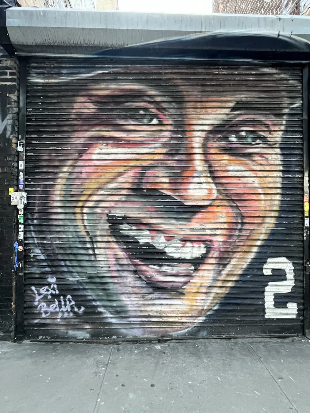 Colorful mural on a rolling metal shutter depicting a smiling person's face in warm tones of orange, yellow, and red with a number 2 in the corner.