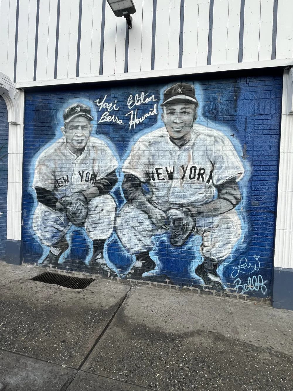 Monochromatic mural on a blue metal shutter showing two vintage baseball players in New York uniforms in classic poses, rendered in white and gray.
