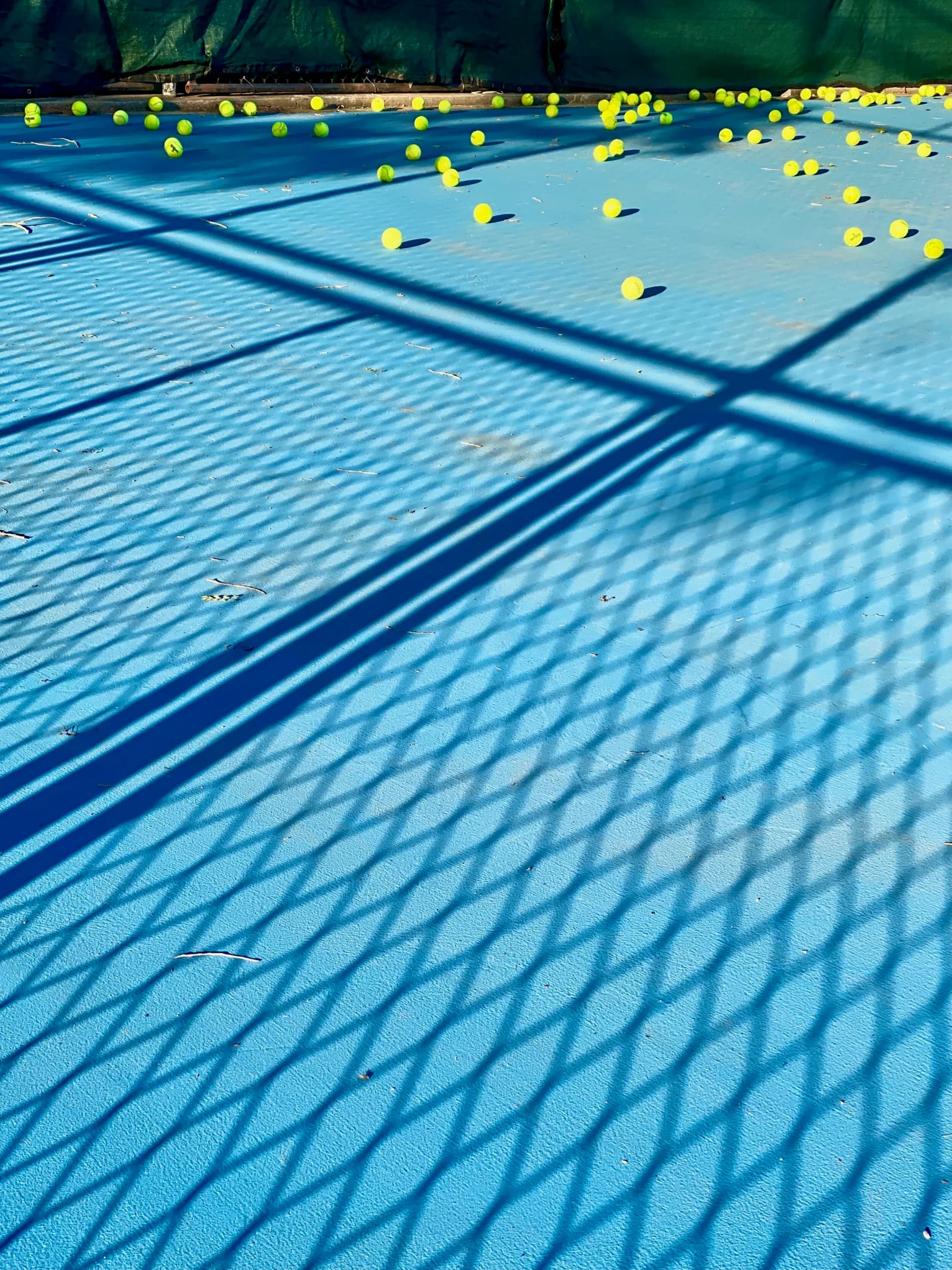 Tennis court photograph with yellow balls scattered across blue court, dramatic net shadows in foreground.