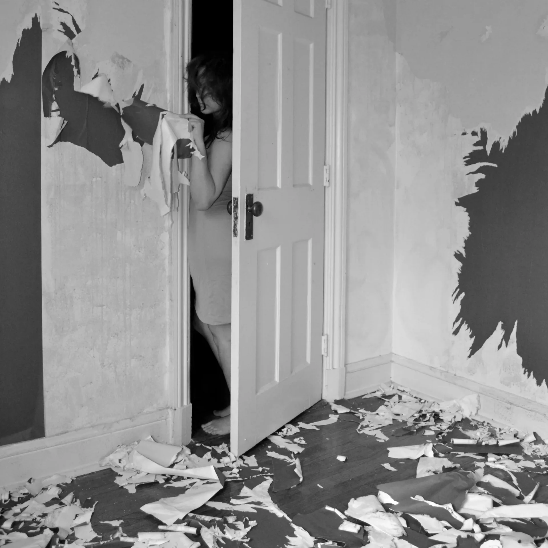 Figure emerging through doorway in room with torn wallpaper and debris on floor