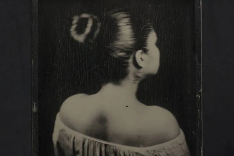 Black and white photograph of a woman from behind, bare-shouldered, with styled hair and dramatic lighting creating strong contrast.
