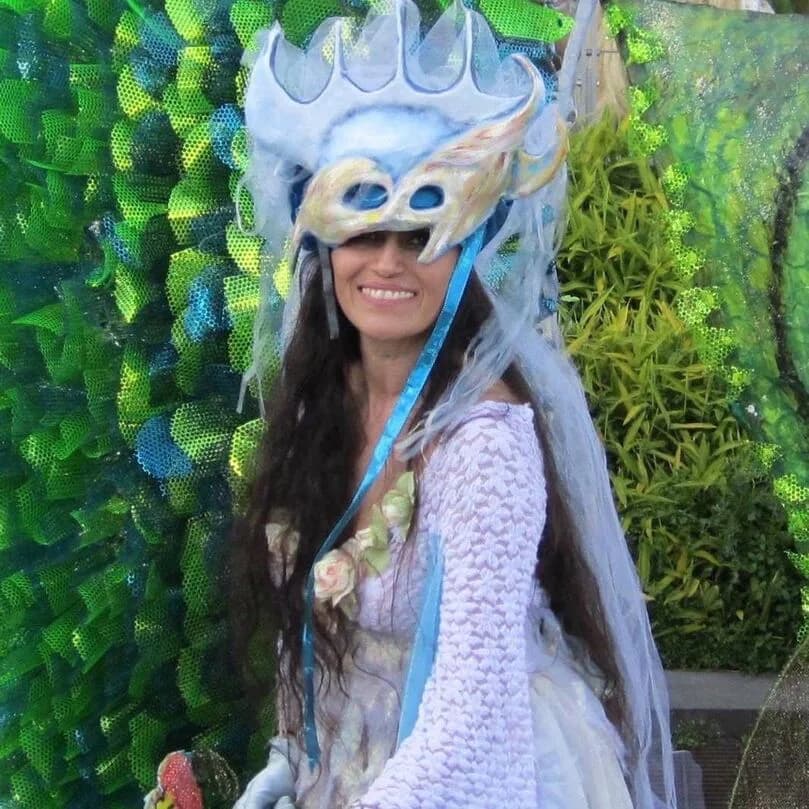 Person wearing an elaborate sculptural mask headdress with white and blue coloring, large eye openings, and flowing fabric elements against vibrant green decorative background.
