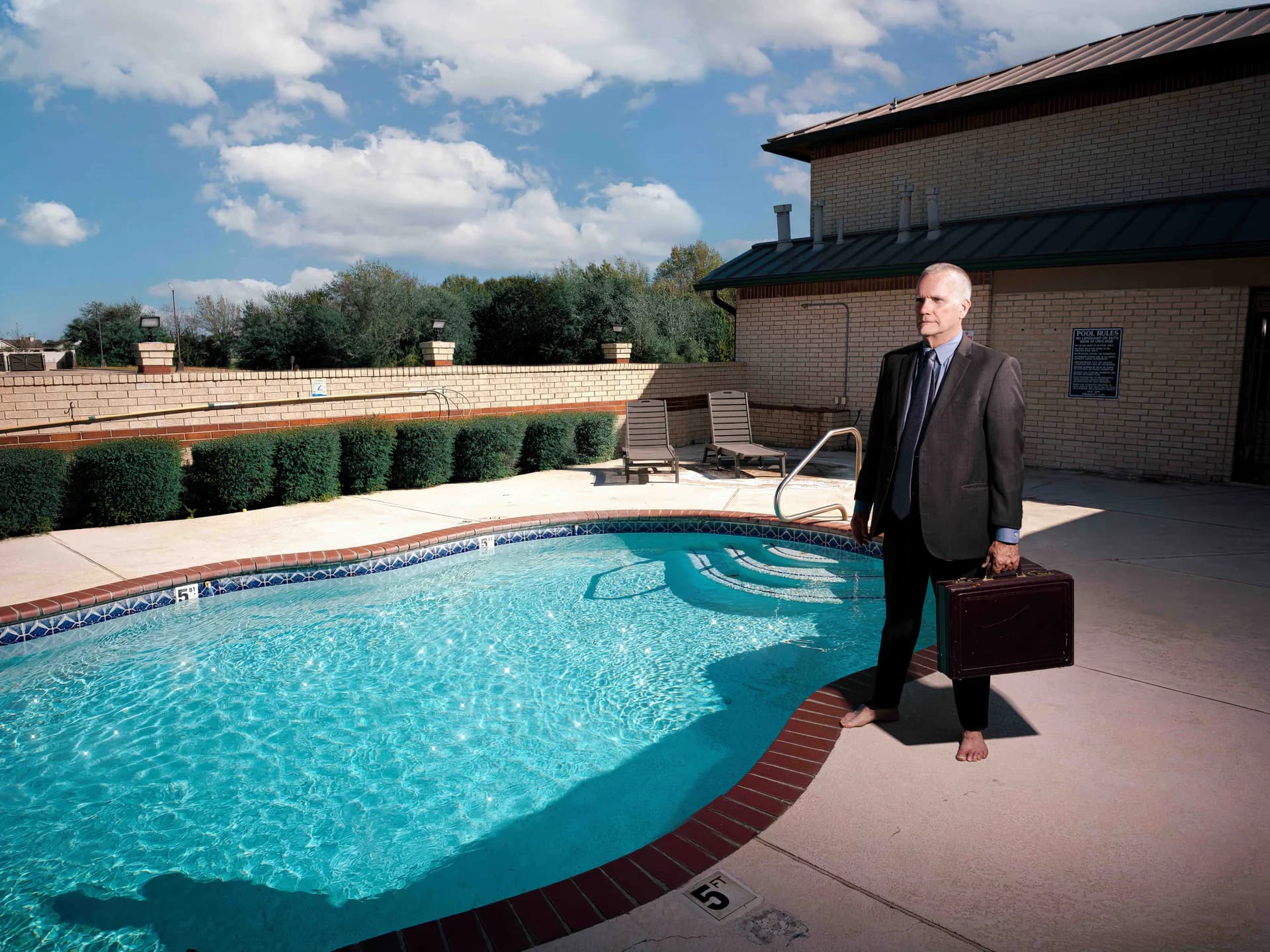 Man in suit standing barefoot poolside with briefcase — surreal editorial photograph
