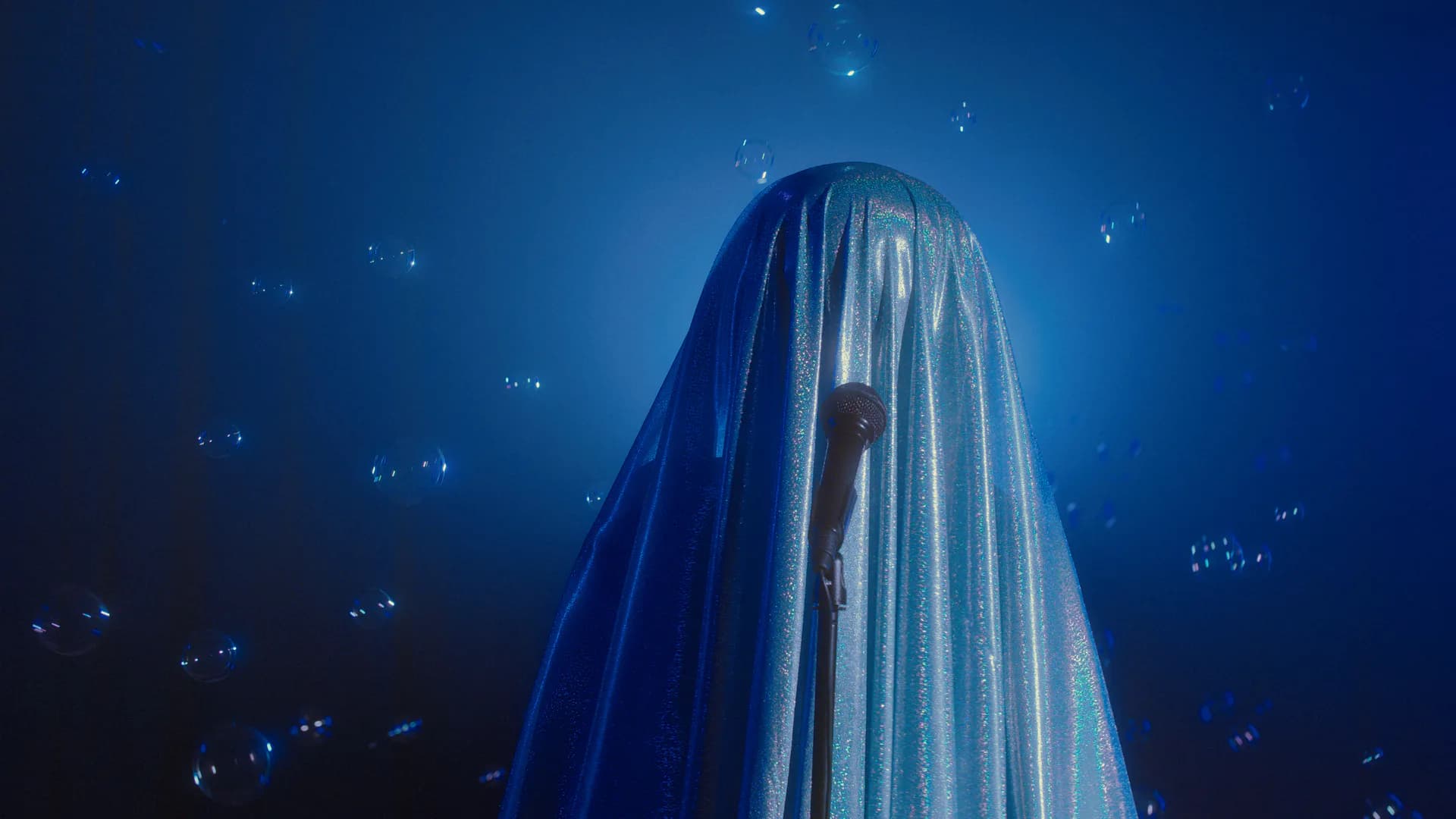 Figure draped in iridescent blue fabric with microphone stand amid floating bubbles