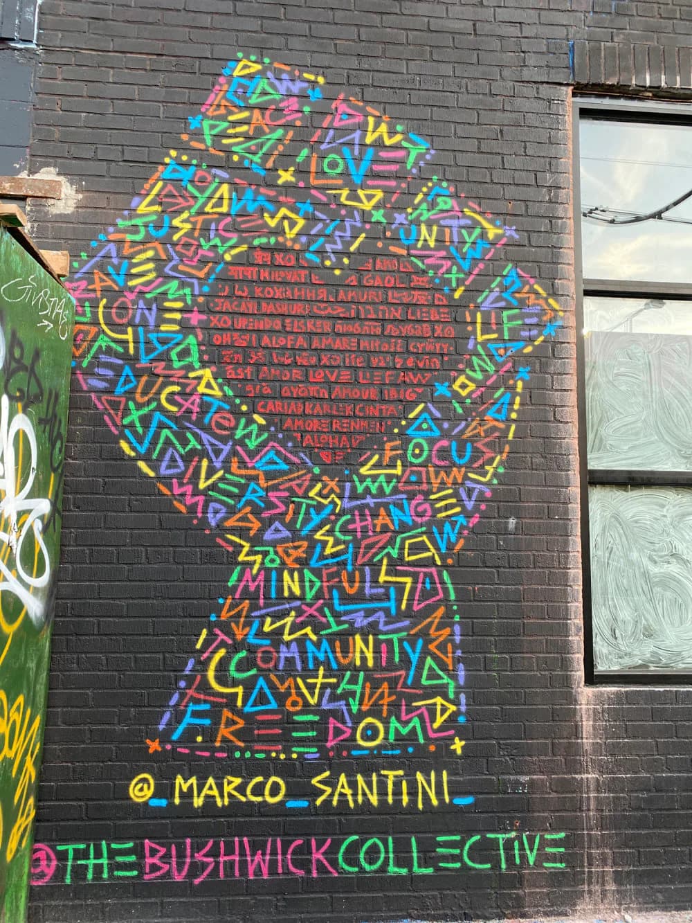 Mural of a raised fist made of colorful hand-lettered words such as Peace, Love, Unity, and Freedom, painted on a brick wall, signed Marco Santini for The Bushwick Collective.