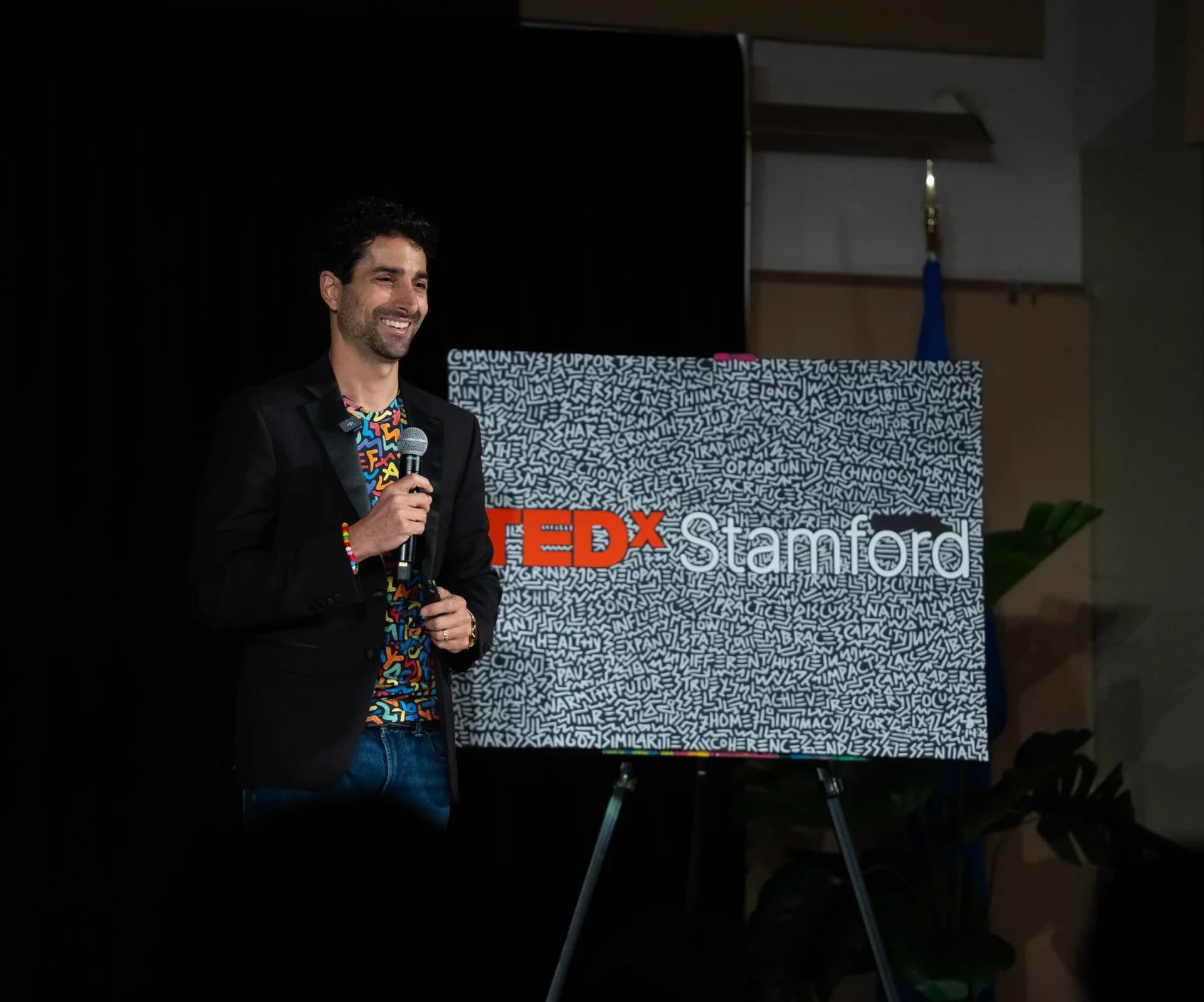 Man in a black blazer holding a microphone and smiling while presenting on a TEDx Stamford stage next to a sign covered in dense typographic patterns.