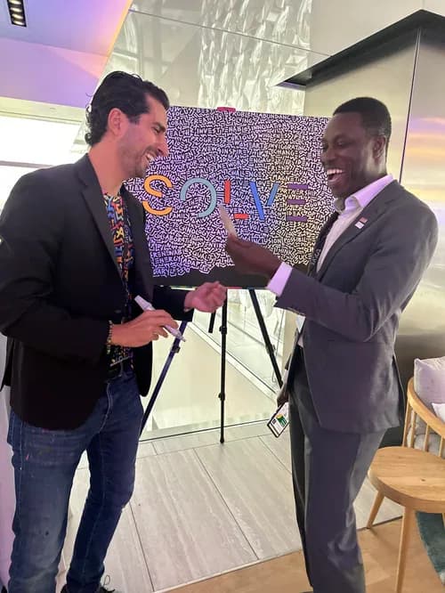 Two men, one in a black blazer and one in a gray suit, laughing together indoors near a colorful typographic artwork reading SOLVE on an easel.