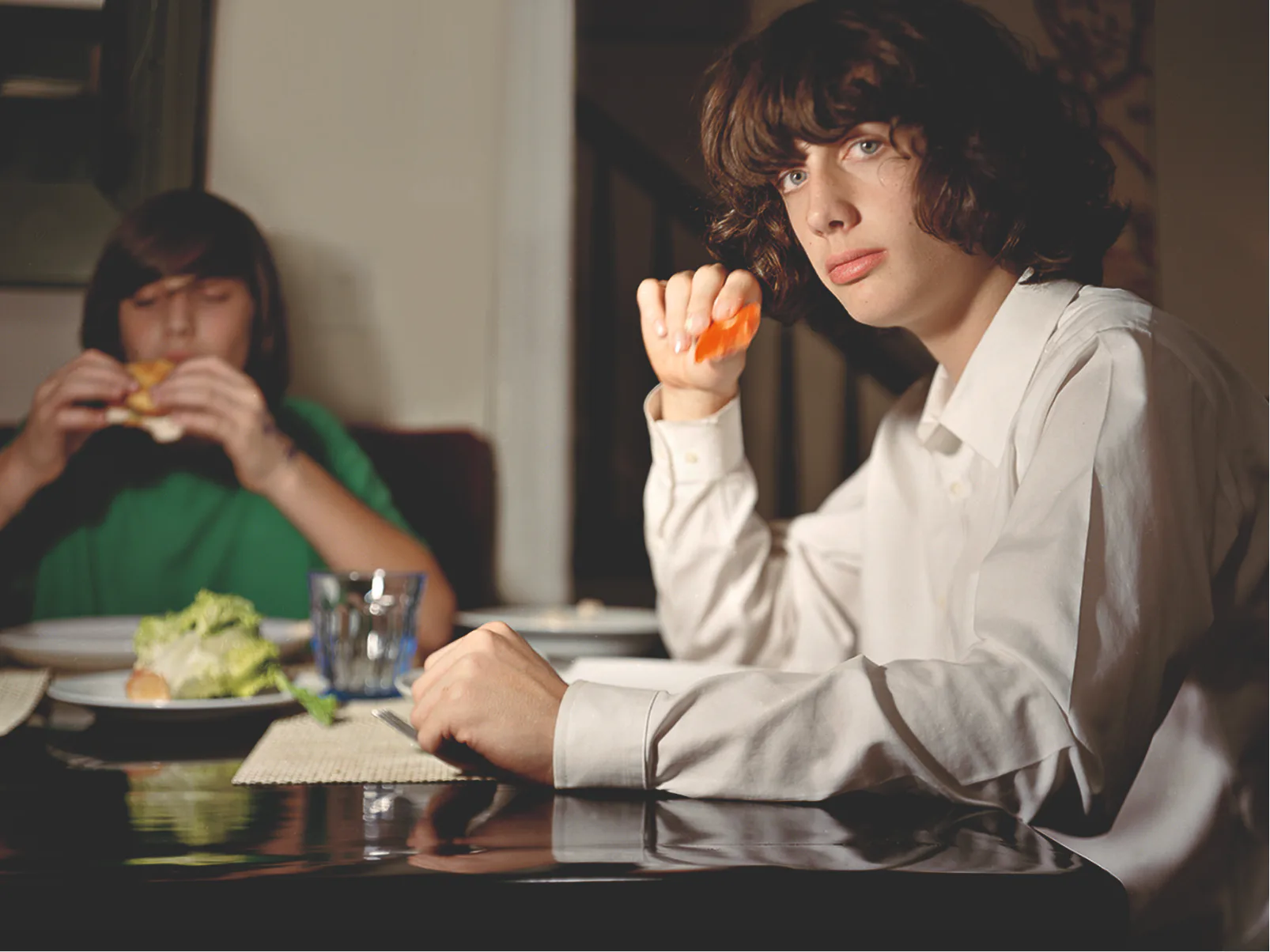 Young man in white shirt eating an orange at a table indoors, with another person eating in the background, warm indoor lighting.