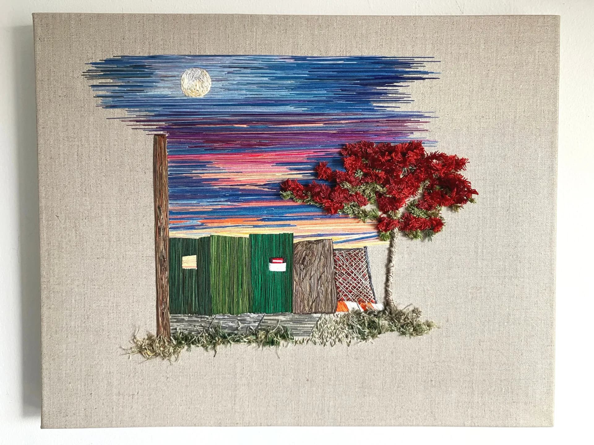 Embroidered textile art on linen showing a moonlit cabin with red tree, starry sky, and stitched landscape elements by Melissa Calderón.
