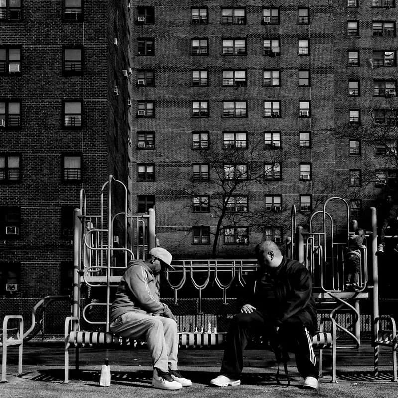 Black and white photograph of two figures sitting on a bench in front of a playground structure with apartment buildings behind.