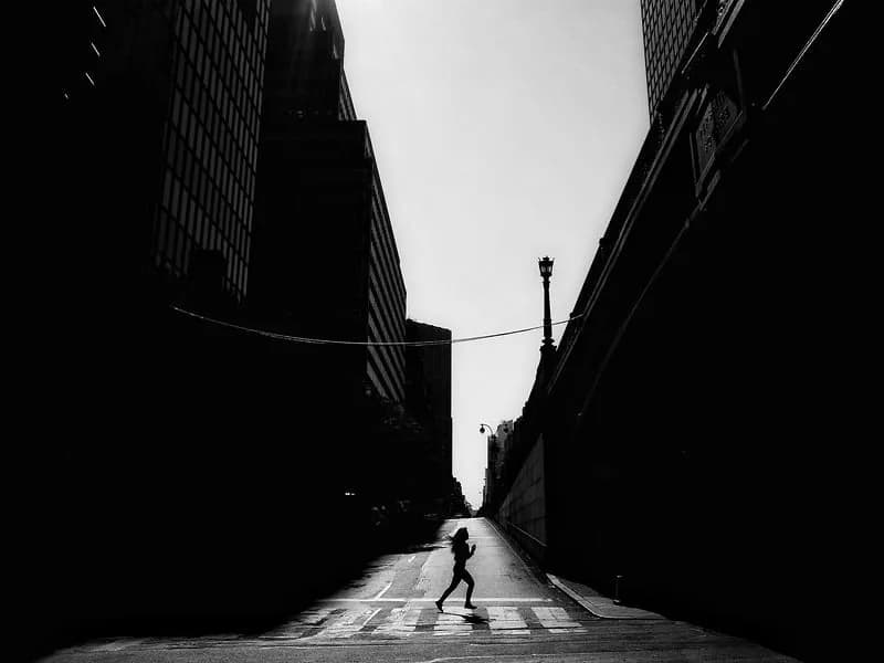 High-contrast black and white photograph of a solitary figure crossing a crosswalk in a narrow urban alley between tall buildings.