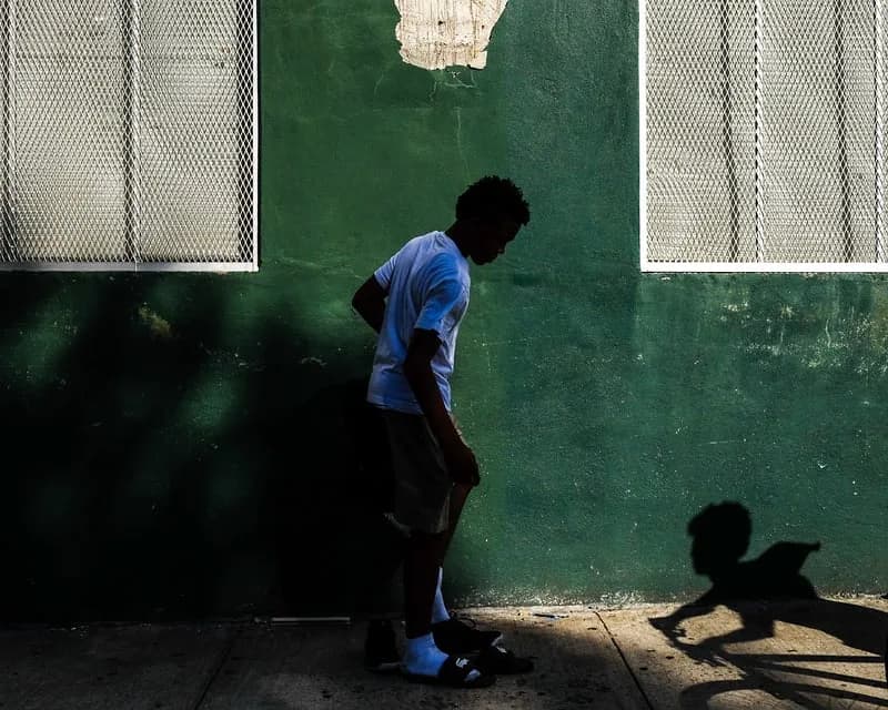 Color photograph of a figure in blue shirt leaning against a weathered green wall with shadow casting across the surface.