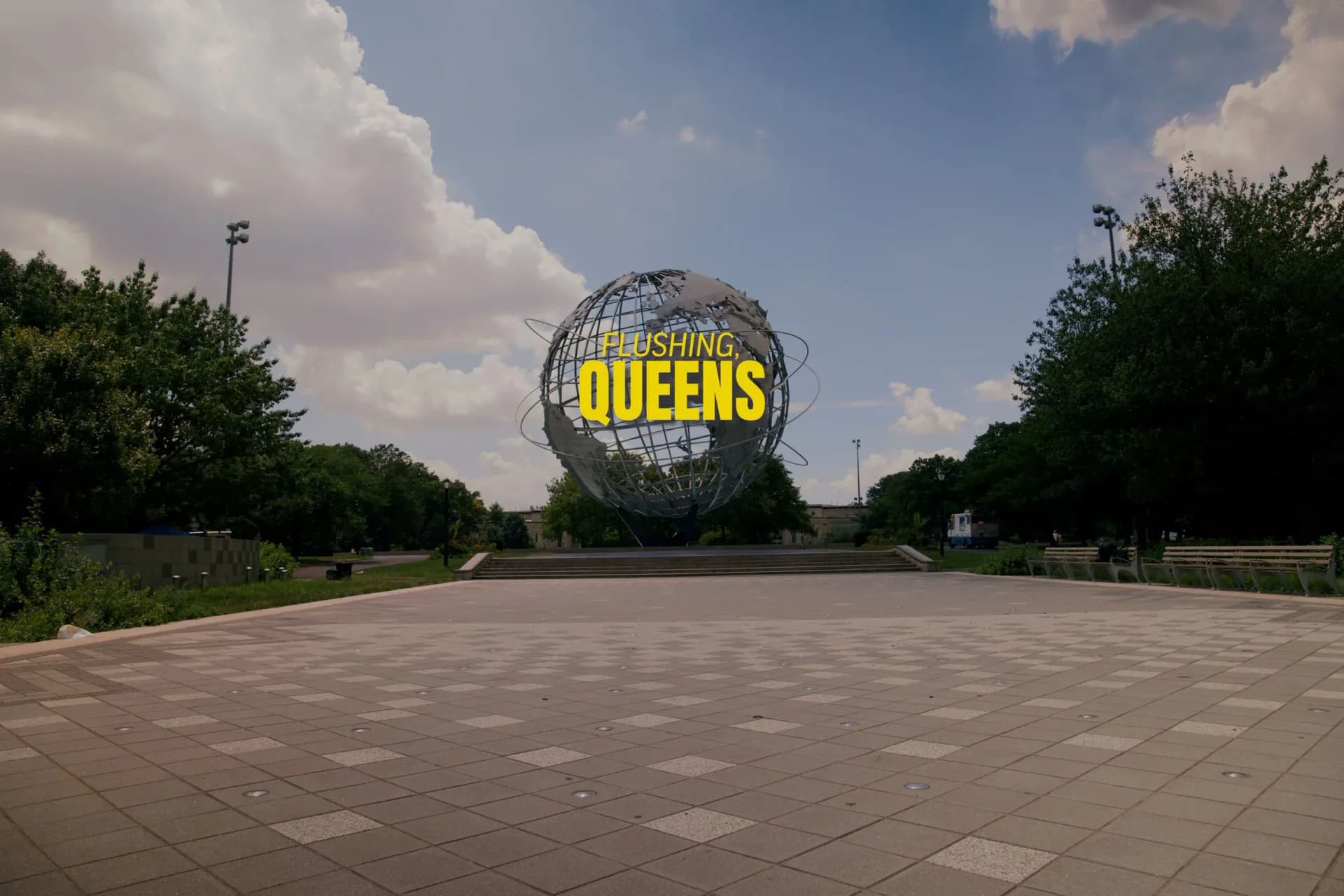 Wide plaza with spherical structure bearing yellow 'FLUSHING QUEENS' text, tree-lined horizon and dramatic sky.