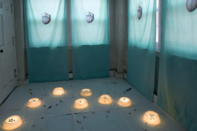 Installation artwork: glowing spherical forms arranged on a floor within turquoise-walled gallery space with overhead lights.
