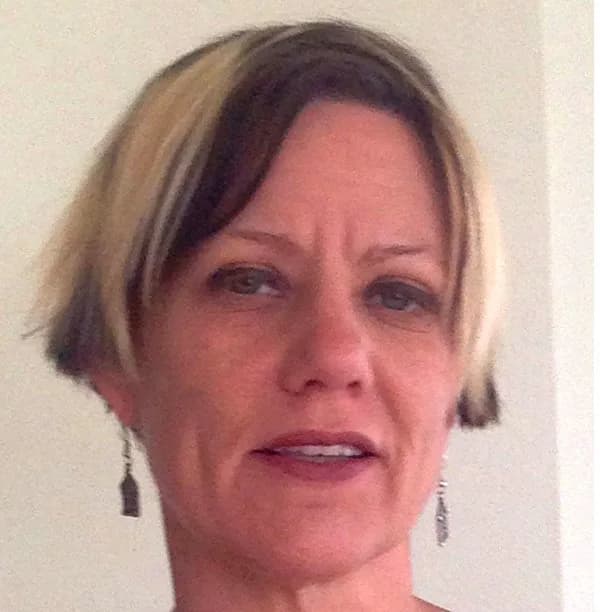 Portrait of a woman with short blonde and dark hair, wearing dangling earrings against a neutral wall.