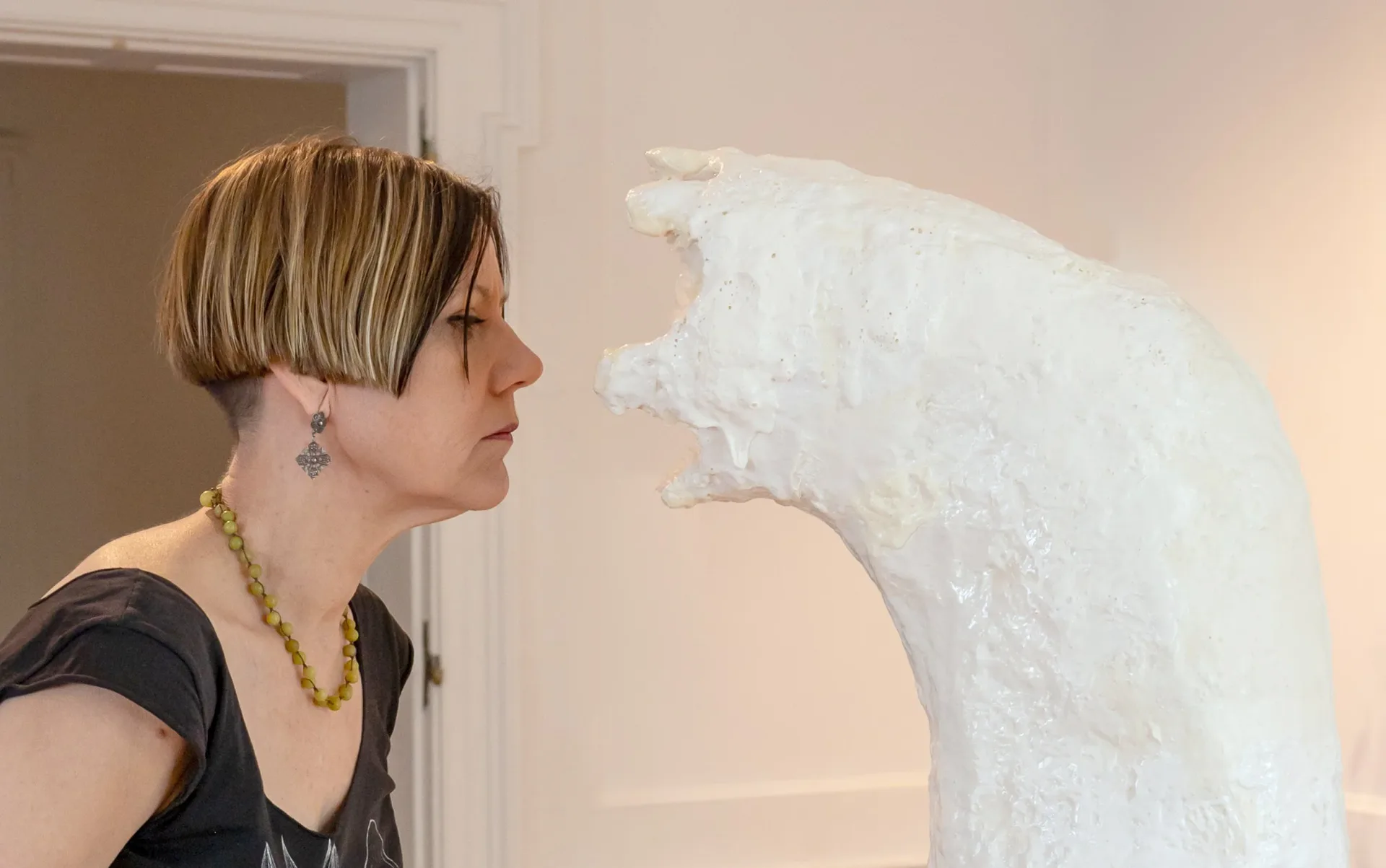 Woman standing in profile next to a large white sculptural form with organic, textured surface and an open circular mouth-like opening.