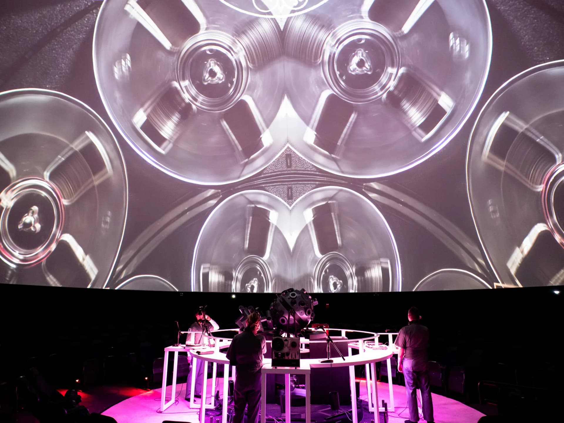Wide view of immersive performance space with performers at tables beneath dome projection of reel-to-reel tape imagery