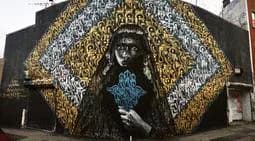 Mural depicting figure in dark clothing with ornate gold circular halo against dark background, holding turquoise object.