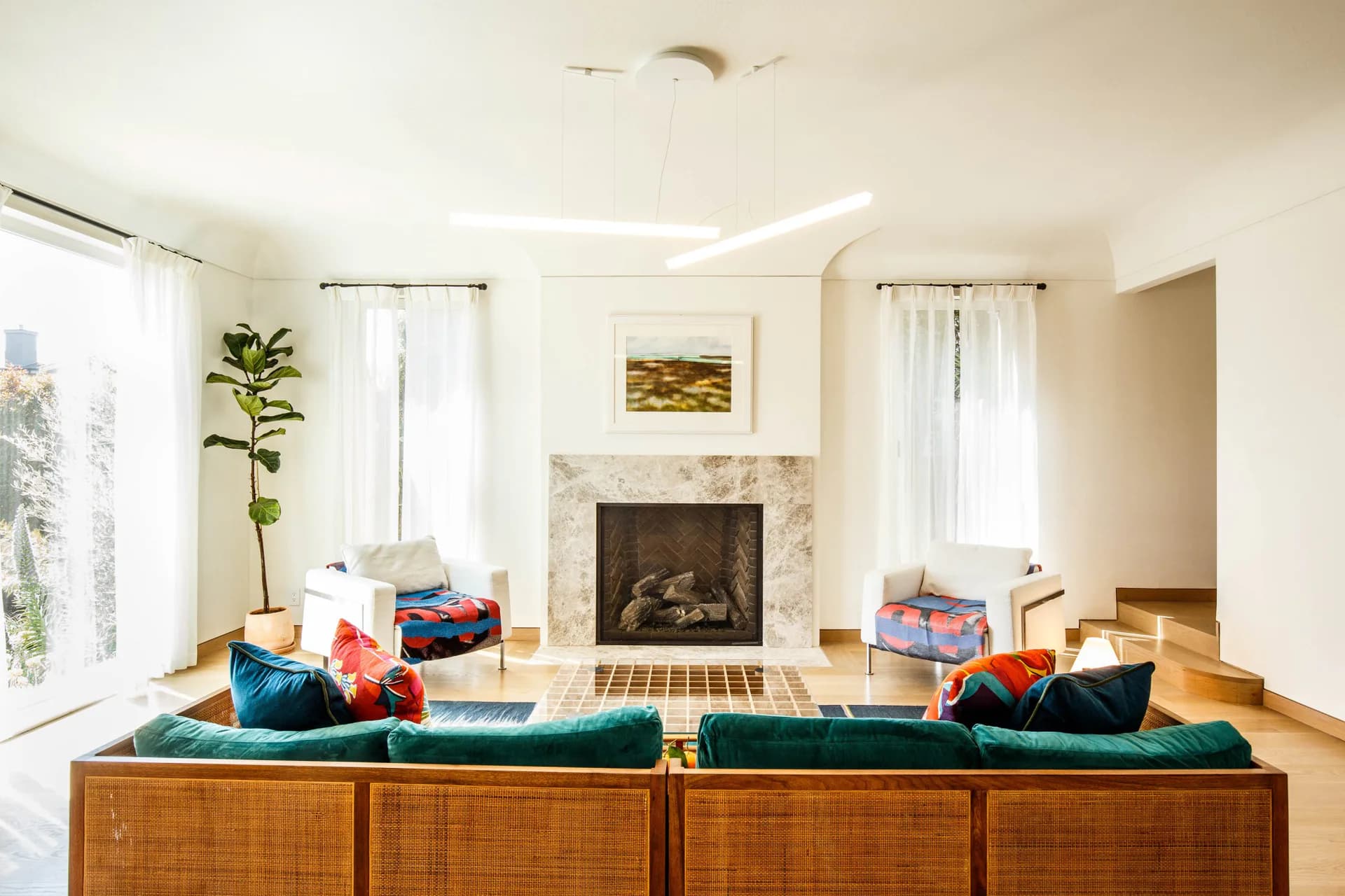 Bright interior living room with teal velvet sofa, fireplace, floor-to-ceiling windows, wooden flooring, and colorful storage boxes.