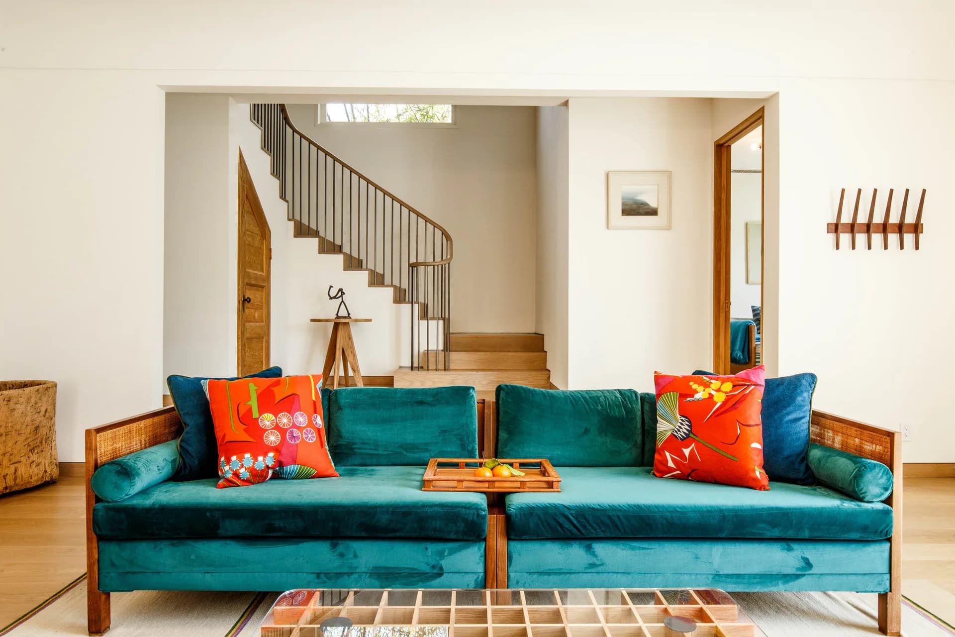 Modern open-plan interior with teal velvet sofa, red and orange patterned pillows, wooden staircase visible through archway, and geometric area rug.